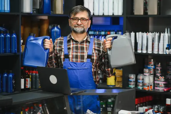 Oto yedek parça mağazasında müşterilere yardım ederken iki kutu motor yağı tutan tamirci, çeşitli otomotiv ürünleri sergiliyor