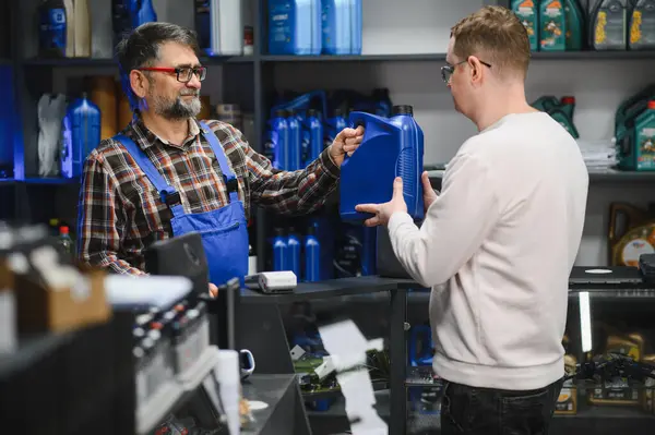 Tamirci oto-gereç dükkanındaki müşterilere bir şişe motor yağı veriyor. Raflarda çeşitli araba bakım ürünleri bulunuyor.