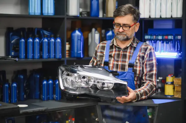 Tecrübeli oto tamircisi, çeşitli araba bakım ürünleriyle dolu raflarla çevrili bir oto yedek parça mağazasında yeni bir far inceliyor.