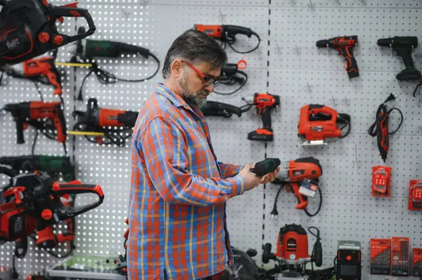 Sakallı, olgun bir adam, çeşitli elektrikli aletler ve ekipmanlarla çevrili, hareketli bir hırdavatçıdan kablosuz bir tornavida seçiyor.