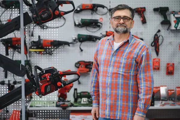 Çeşitli elektrikli testereler ve elektrikli testerelerle çevrili bir hırdavatçıda duran kendinden emin bir satıcının portresi müşterilere yardım etmeye hazır.