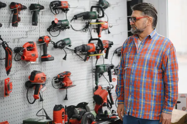 Hırdavatçıda duvara asılı elektrikli aletlere bakan kıdemli satıcı, müşteri için en iyi seçeneği seçiyor