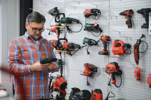 Sakallı olgun müşteri bir hırdavatçıda elektrikli bir araç seçiyor, kutunun üzerindeki özellikleri okuyor.