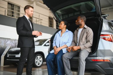 Araba satıcısı, araba galerisindeki açık bagajda oturan Afro-Amerikan çifte araba özelliklerini açıklıyor.