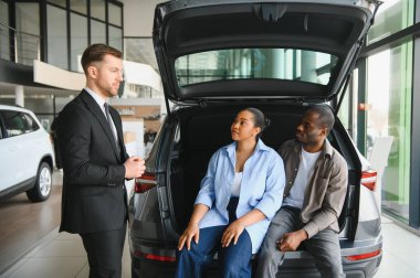 Araba satıcısı, yeni bir aracın bagaj boşluğunu ön plana çıkarırken, aynı zamanda çok hareketli bir bayilik ortamında bir çiftle karşılaşmış.