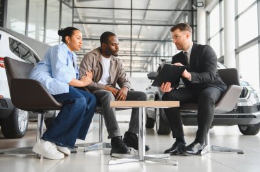 Satış elemanı, modern bir araba galerisinde finansal seçenekleri veya araç özelliklerini tartışan bir çifte kontrat gösteriyor.