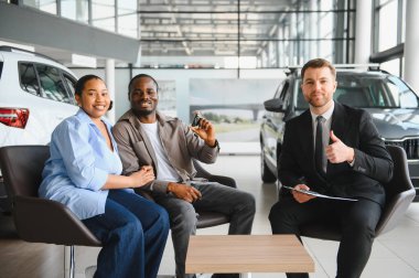 Araba anahtarlarını tutan mutlu çift yeni alımlarını gösteriyor. Satıcıyla oturuyorlar ve bayilikte başparmak kaldırıyorlar.