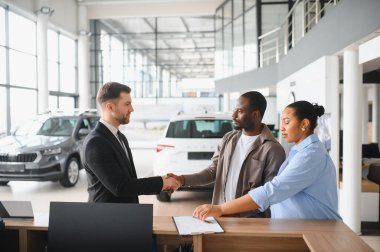 Modern bir galeriden yeni bir araba alırken profesyonel araba satıcısıyla tokalaşan mutlu çift.