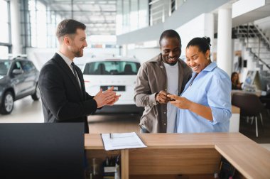 Gülümseyen Afrikalı Amerikalı çift telefon kullanıyor, galeride yeni bir araba seçiyor, satıcıyla konuşuyor.