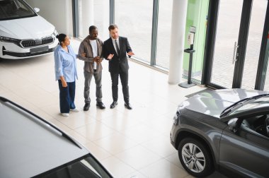 Araba satıcısı, modern bir bayiliğin içinde Afro-Amerikan bir çifte yeni bir araba sunuyor. Araç alımı sırasında yardım ve rehberlik öneriyor.