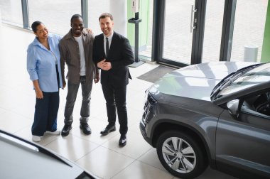 Mutlu çift, yeni arabalarını modern bir bayilikte seçerken profesyonel bir satıcıyla takılıyor, neşe ve heyecan saçıyor.