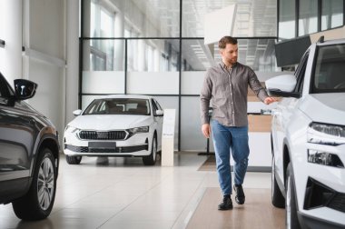 Otomobil galerisinde yeni bir beyaz arabayı inceleyen bir adam satın alma kararı vermeden önce özelliklerini ve kalitesini dikkatlice değerlendiriyor.