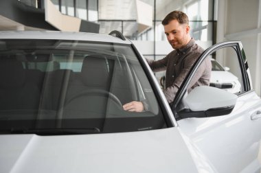 Yeni bir arabanın içini inceleyen adam, bir araba galerisinde modern vitrinlerle çevriliyken direksiyonu hisseden adam.