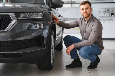 Bir araba galerisinde çömelmiş ve parlak gri bir SUV 'u teftiş eden bir adam, satın alma kararını düşünürken özelliklerini değerlendirir.