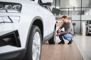 Gösterişli beyaz bir arabanın yanında diz çökmüş, parlak, çağdaş bir sergi salonunda özelliklerini dikkatle inceliyor.