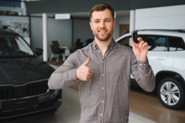 Gülümseyen adam gururla araba anahtarlarını gösteriyor ve bir araba galerisinden aldığı son alımdan duyduğu memnuniyeti dile getiriyor.