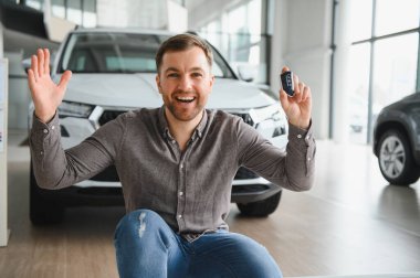 Heyecanlı müşteri gururla yeni araba anahtarlarını sergiliyor. Parlak ve modern bir araba galerisinde başarılı bir alımı kutluyorlar.