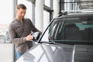 Modern bir araba galerisinde yeni bir araç seçerken yan aynayı ayarlayan adam güven ve memnuniyet gösterisi yapıyor.