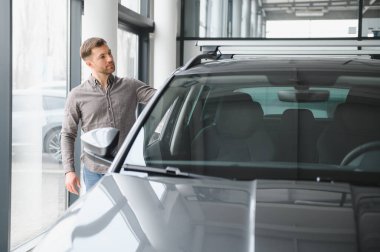 Otomobil galerisinde yeni bir arabayı inceleyen, çeşitli satın alma seçeneklerini değerlendiren, özellikleri ve stilleri değerlendiren bir adam.