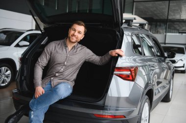 Açık araba bagajında oturan mutlu müşteri, araba galerisindeki yeni aracın özelliklerini inceliyor.