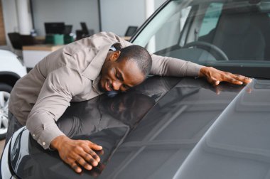 Heyecanlı bir Afrikalı Amerikalı adam yeni arabasını bir araba galerisinde kucaklıyor ve satın almasından duyduğu neşe ve memnuniyeti dile getiriyor.