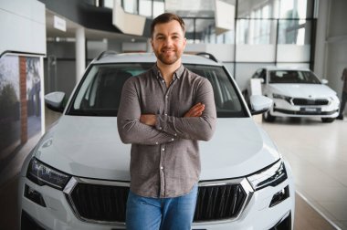 Yeni beyaz bir arabanın önünde kolları çapraz duran mutlu müşteri yeni aracını bir araba galerisinde seçiyor.