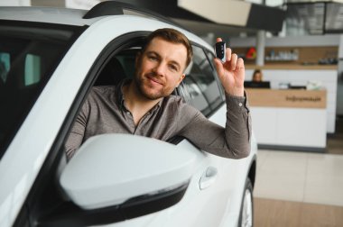 Gülümseyen adam galeride yeni aldığı aracında otururken yeni araba anahtarını gösteriyor.