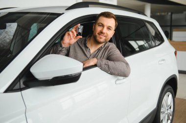 Gülümseyen adam, galeride yeni bir araçta otururken arabanın anahtarlarını tutuyor. Mutlu bir müşteri olarak gurur ve heyecan saçıyor.