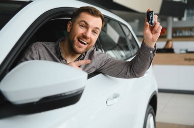 Yeni araba anahtarlarını gösteren mutlu adam, son alımını kutlamak için galerinin içindeki araca işaret ediyor.