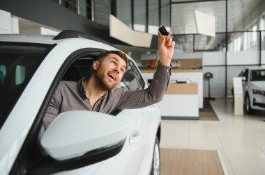 Hevesli bir adam, elinde anahtarlar, modern bir araba galerisinde yeni bir araç almanın heyecanını simgeliyor.