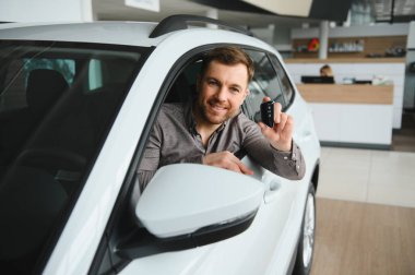 Gülümseyen bir adam yeni bir arabada oturuyor, elinde anahtar var, modern bir araba galerisinde, bir araç satın almanın heyecanını sergiliyor.