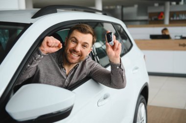 Yeni araba alımını kutlayan heyecanlı adam, modern araçlarla dolu canlı bir galeride arabanın anahtarlarını neşeyle tutuyor.