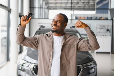 Yeni araba alımını kutlayan heyecanlı adam modern araçlarla dolu canlı bir galeride araba anahtarlarını gururla sergiliyor.