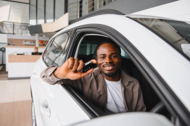 Gülümseyen adam elinde araba anahtarlarıyla galeride yeni bir araçta otururken mutluluk ve memnuniyet saçıyor.