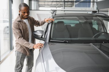 Afro-Amerikalı bir adam araba galerisinde yeni gri bir arabayı teftiş ediyor, kaputa dokunuyor ve çatı rafını kontrol ediyor.