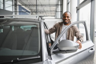 Gülümseyen Afrikalı Amerikalı bir adam galeriden araba alma tecrübesinden zevk alıyor, yeni aracına mutlulukla biniyor.