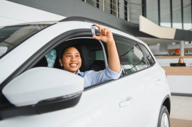 Bir araba galerisinde yeni arabasının içinde oturan heyecanlı müşteri, araba anahtarlarını gururla tutuyor ve neşe ve başarıyla gülümsüyor.