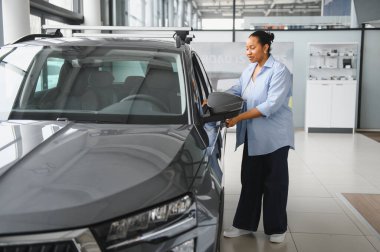 Afro-Amerikalı bir kadın galeride yeni bir araba seçerken yan aynayı ayarlıyor.
