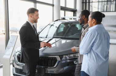 Araba satıcısı, modern bir galeride, özellikleri ve alım opsiyonlarını tartışan bir çifte yeni bir araba sunuyor.