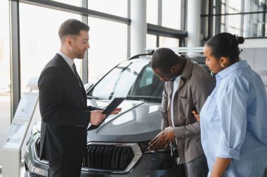 Araba satıcısı Afrikalı Amerikalı çifte yeni bir araç gösteriyor. Modern galeride özellikleri ve alım opsiyonlarını tartışıyorlar.