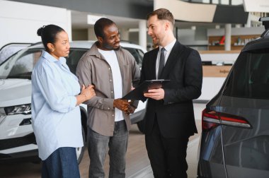 Araba satıcısı elinde bir not panosuyla modern bir otomobil galerisinde Afro-Amerikan bir çifte yeni bir araba sunuyor. Seçenekleri ve anlaşmaları tartışıyorlar.