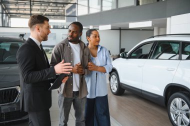 Araba satıcısı, modern bir galeride, özellikleri ve alım opsiyonlarını tartışan bir çifte yeni bir araba sunuyor.