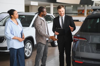 Afro-Amerikan çifte yeni otomobil sunan profesyonel araba satıcısı modern bayilikte alım opsiyonlarını tartışıyor.
