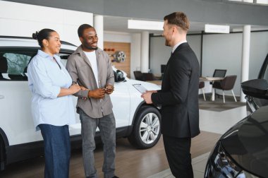 Gülümseyen araba satıcısı elinde not panosuyla modern bayilikteki Afrikalı Amerikalı çifte yeni bir araba gösteriyor.