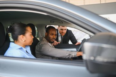 Araba satıcısı, yeni bir araba almak isteyen Afrikalı Amerikalı bir çifte yeni bir arabanın özelliklerini gösteriyor.