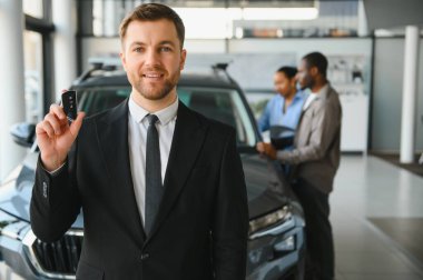 Araba satıcısı gülümsüyor ve elinde araba anahtarı var. Arka planda yeni arabalarını seçen genç bir çift var.