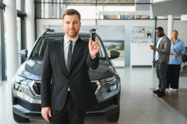 Araba satıcısı heyecanlı bir çiftin araba anahtarını sunuyor. Parlak, modern bir galeride yeni arabalarını seçiyorlar.