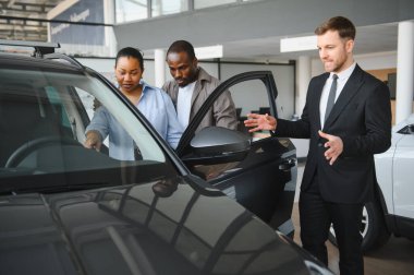 Araba satıcısı bir çiftle takılıyor, modern bir galeride yeni bir arabanın özelliklerini ön plana çıkarıyor, satın almaları konusunda heyecan yaratıyor.