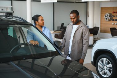 Araba galerisinde Afro-Amerikan bir çifte yeni bir arabanın özelliklerini gösteren satıcı, doğru seçimi yapmalarına yardım ediyor.