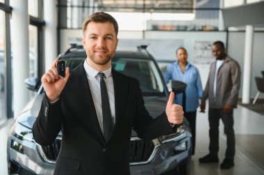 Araba satıcısı araba anahtarını gösteriyor ve yeni arabasının önünde bayide mutlu bir çiftle başparmak işareti yapıyor.